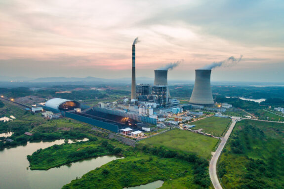 Aerial view shows a nuclear power plant, surrounded by lush green areas and water, with mountains in distance.