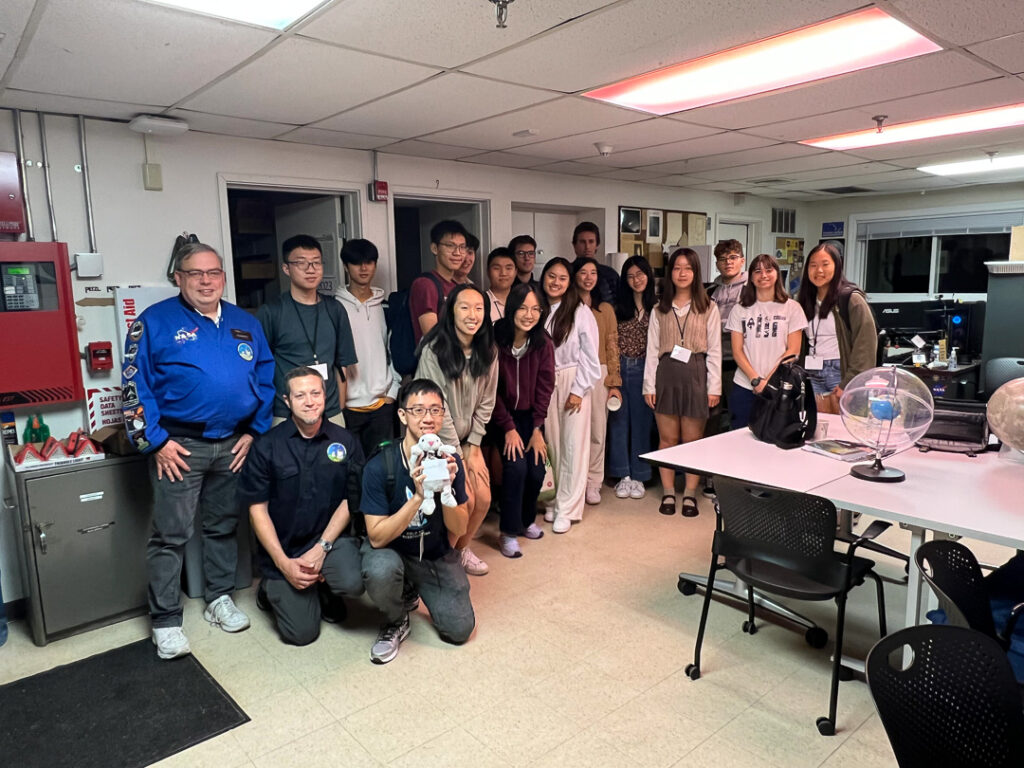 A group photo of 19 people inside the MIT Wallace Observatory in regular lighting.