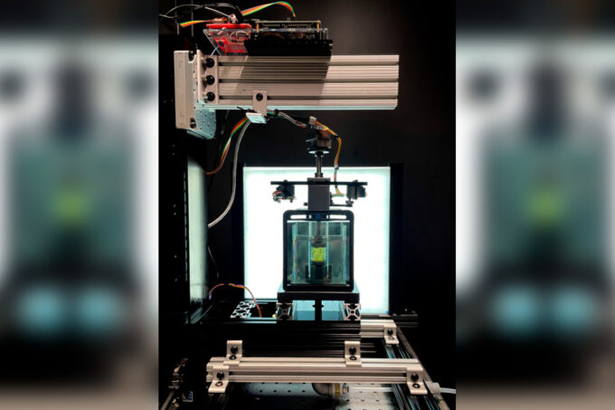 In the lab, the prototype scanner is made of metal and wires, with a sturdy clear box inside. Inside the box is a glass tube with yellow material inside. A lightbox is in the background, providing light.