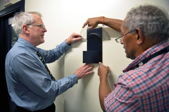 Richard Binzel and Glenn Silva hold a 3D printed plaque depicting Saturn's rings for position on a blank wall.