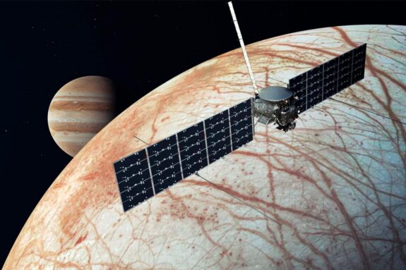 Photo illustration of a spacecraft with large solar panels hovering over a reddish striped moon with planet Jupiter in the background.